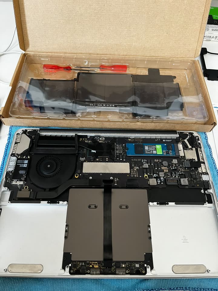 Notebook Apple aberto na bancada, com bateria nova na embalagem ao lado.